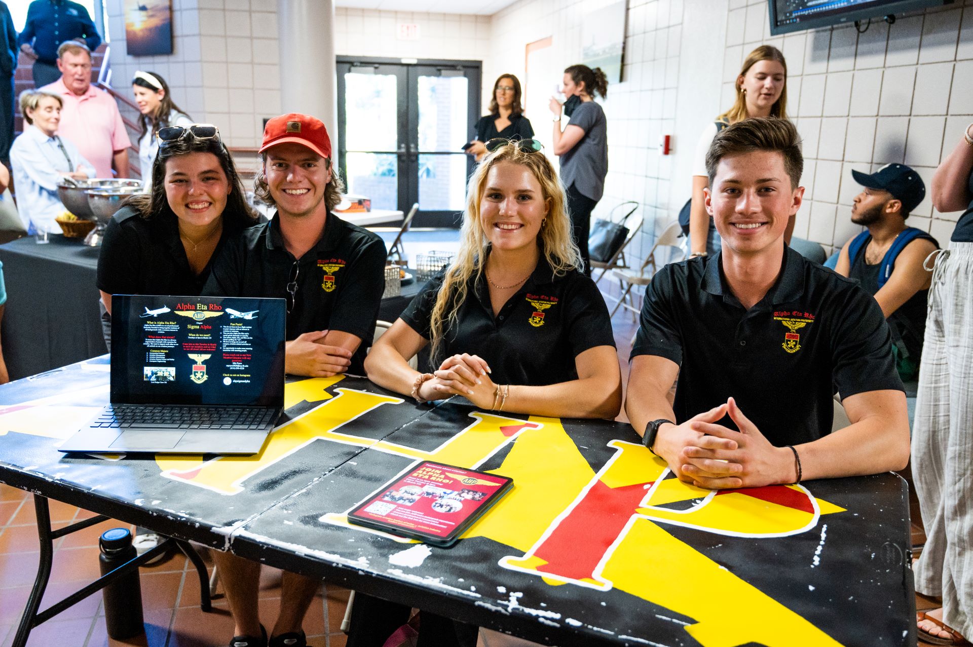 Four smiling students wearing team polo shirts seated at a recruitment table displaying a laptop and informational materials at a university event